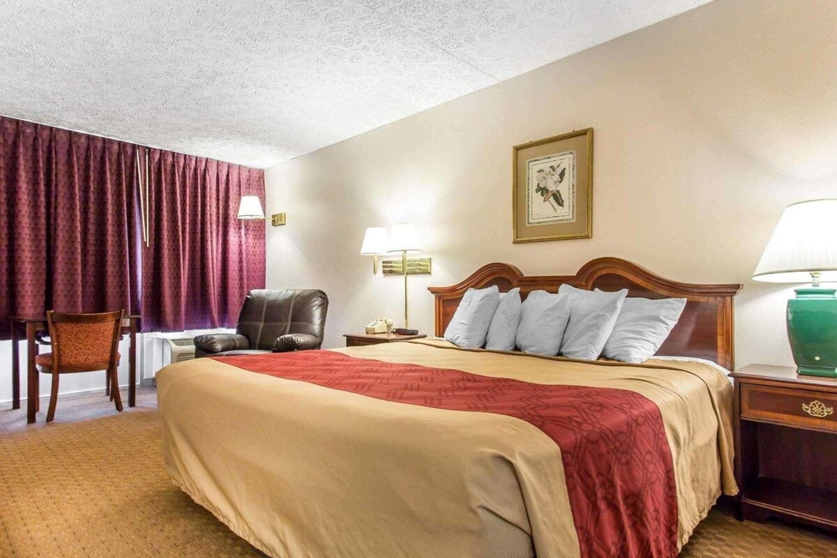 A hotel room with a large bed, a desk, a chair, and a nightstand with a lamp.