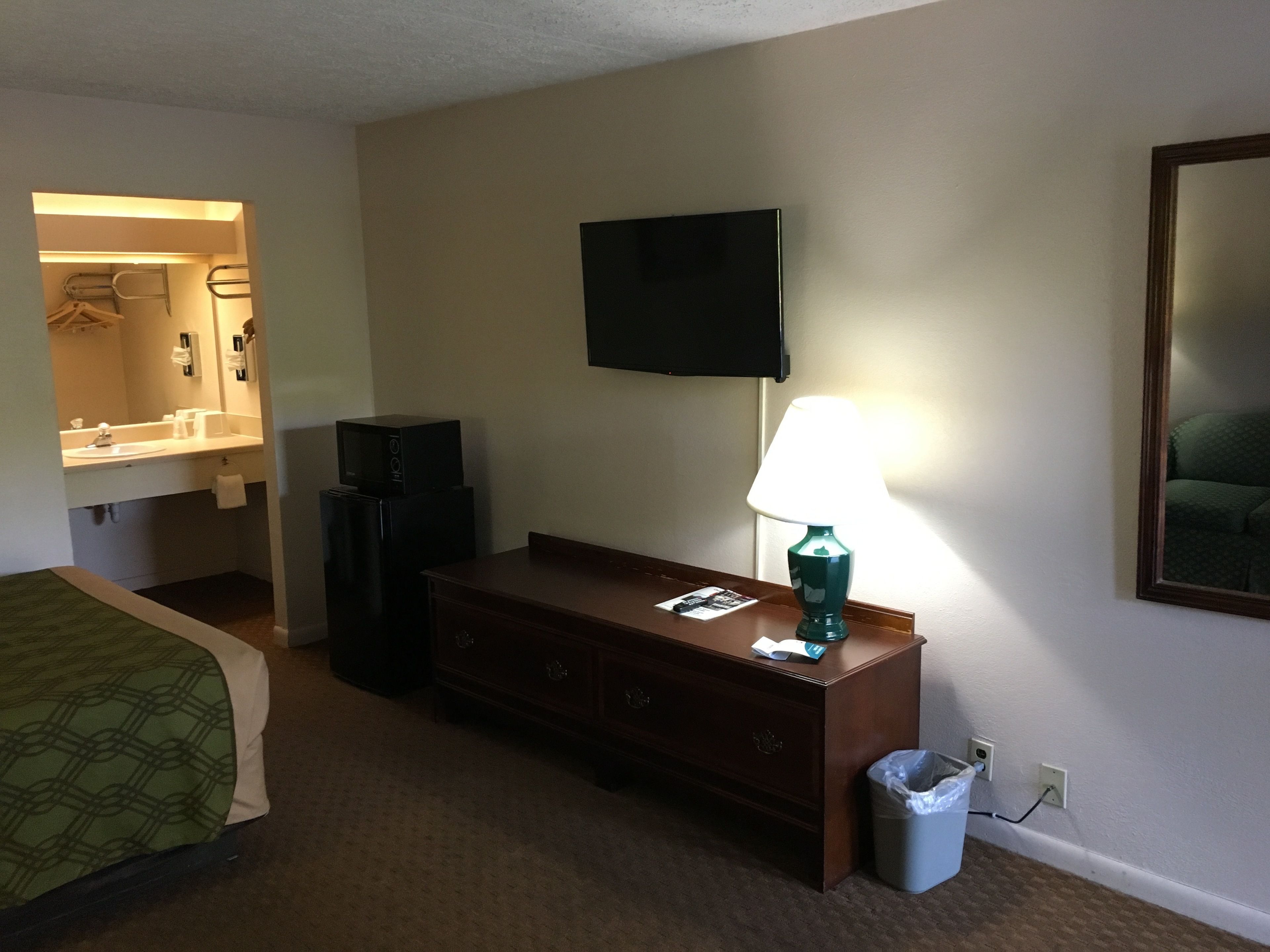 A hotel room with a bed, a dresser, a TV mounted on the wall, and a lamp on the dresser.