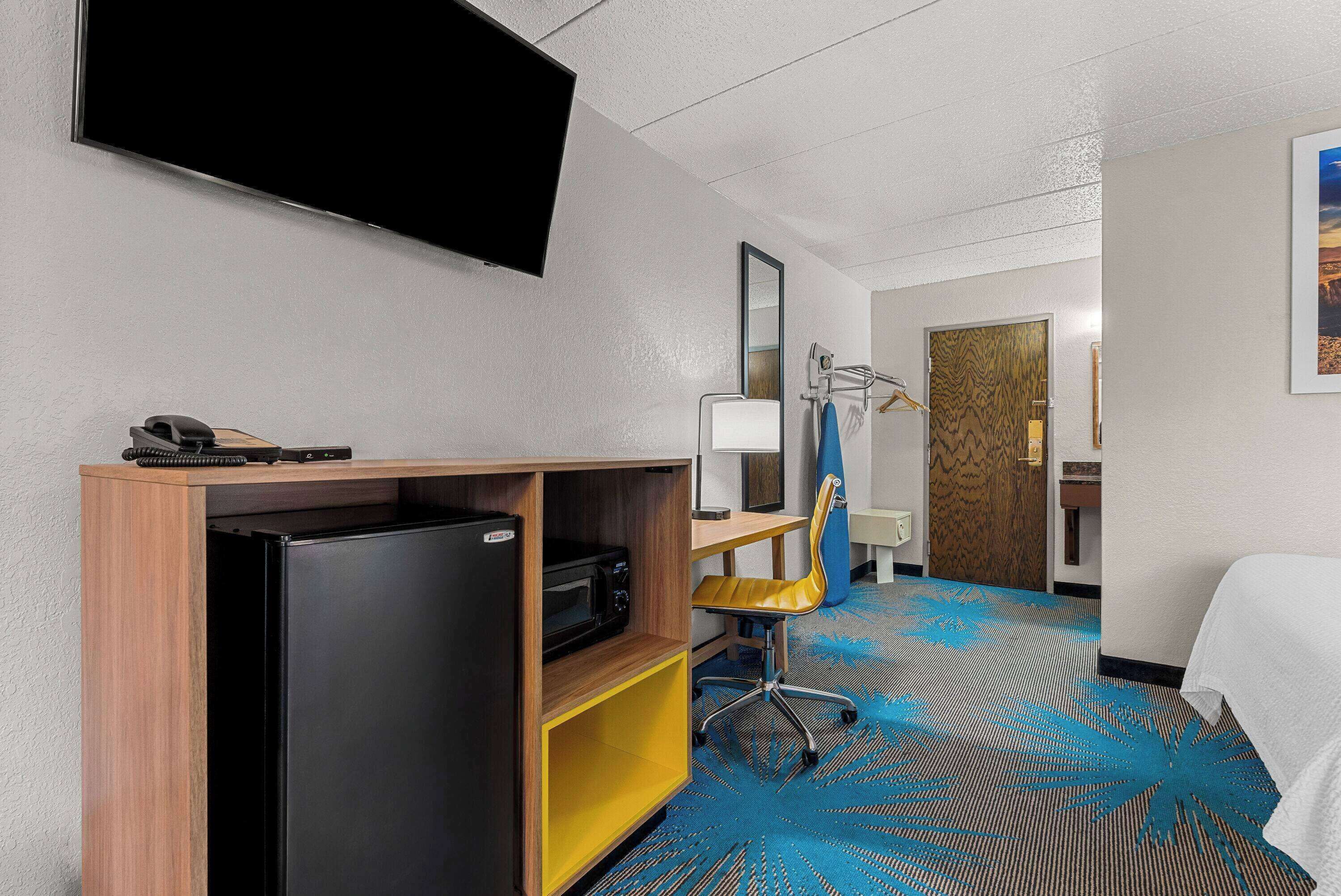 Une chambre d’hôtel comprenant un lit, un bureau avec un ordinateur, une chaise, un réfrigérateur, un four à micro-ondes, un téléphone, un téléviseur à écran plat fixé au mur et une porte donnant sur une autre pièce.