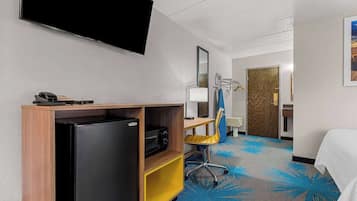Une chambre d’hôtel comprenant un lit, un bureau avec un ordinateur, une chaise, un réfrigérateur, un four à micro-ondes, un téléphone, un téléviseur à écran plat fixé au mur et une porte donnant sur une autre pièce.