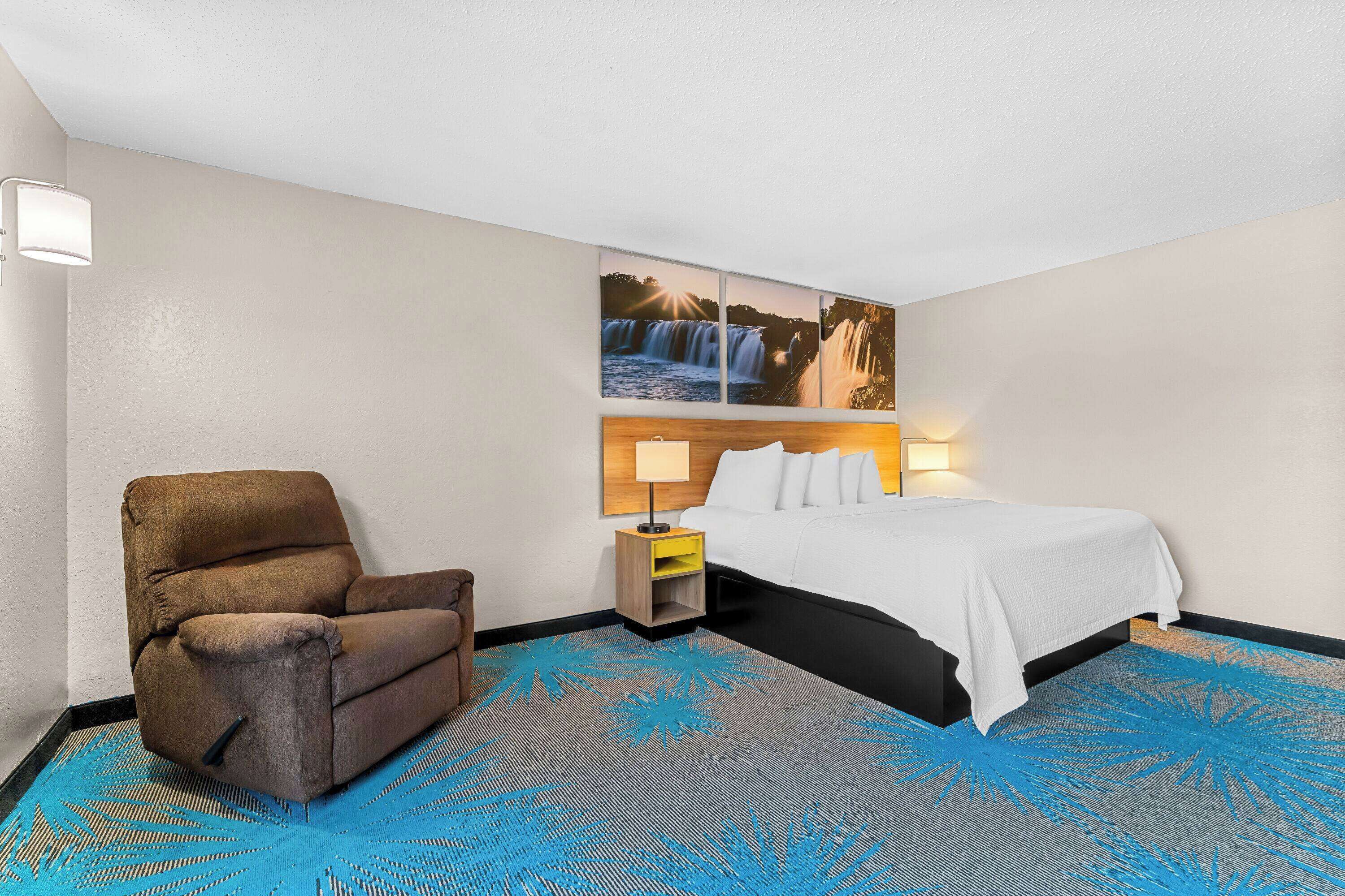 Habitación de hotel con cama, una silla, una mesita de noche y un cuadro colgado en la pared con una cascada.