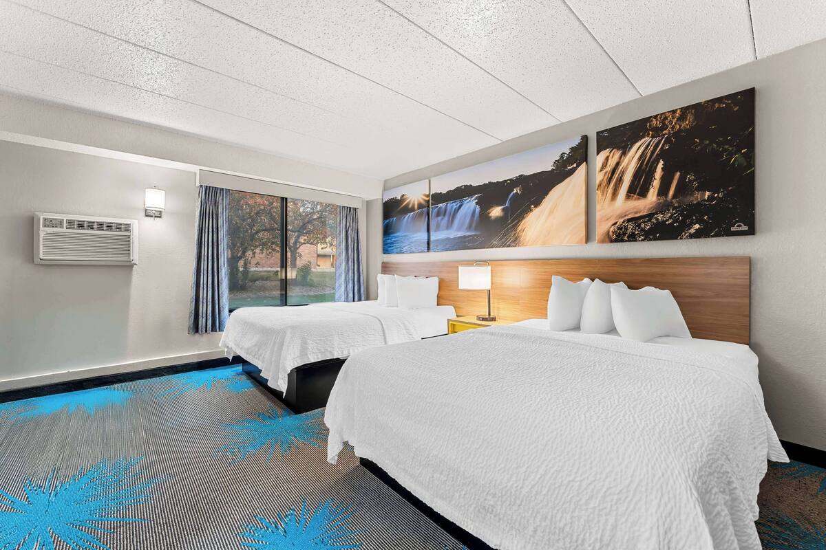 A hotel room with two beds, a large window with curtains, a wall-mounted air conditioner, and a waterfall painting.