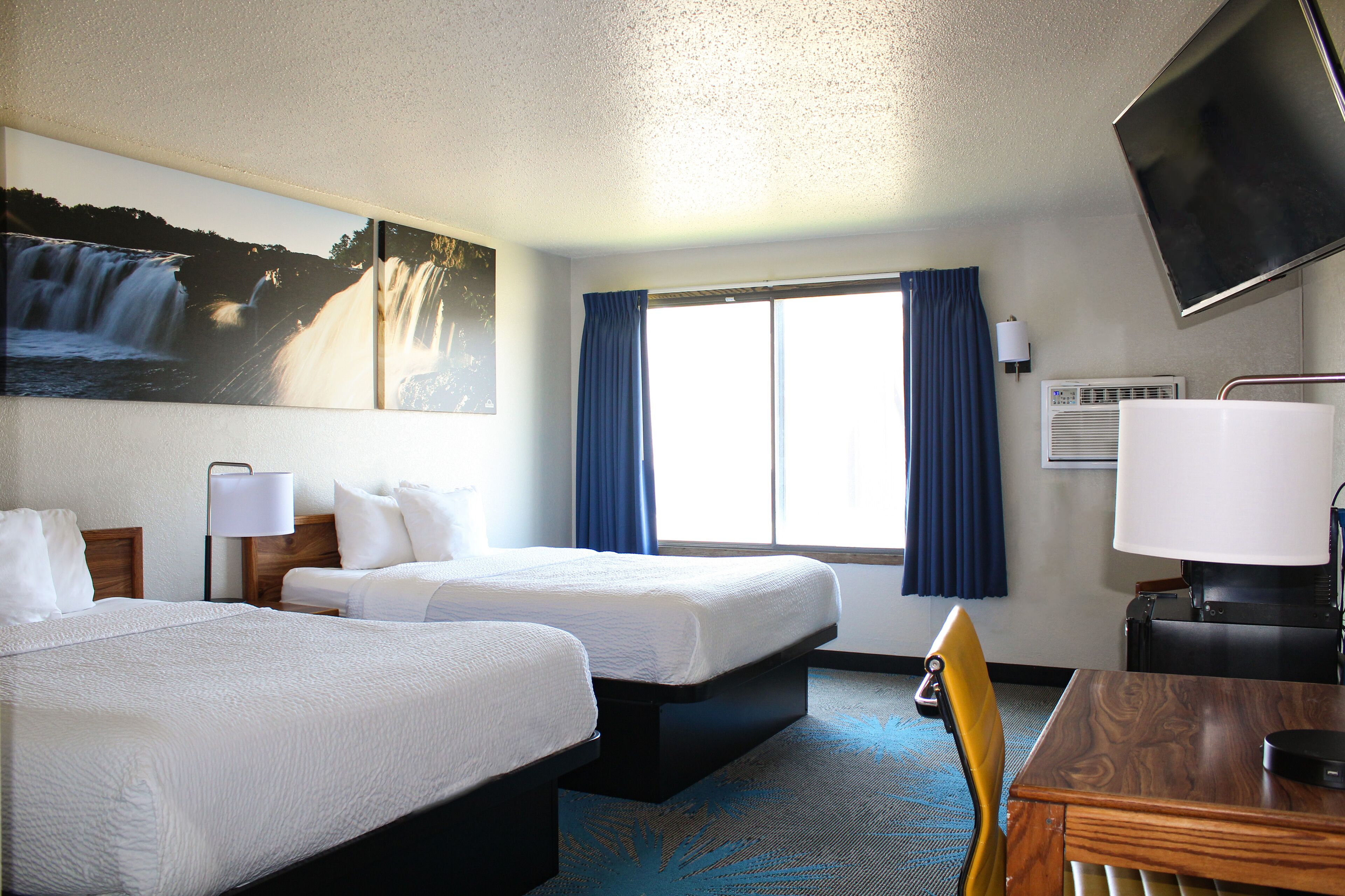 A hotel room with two beds, a desk, a chair, a TV, and a window with curtains.