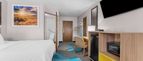 A hotel room with a bed, a desk, a TV, and a bathroom visible through an open door.