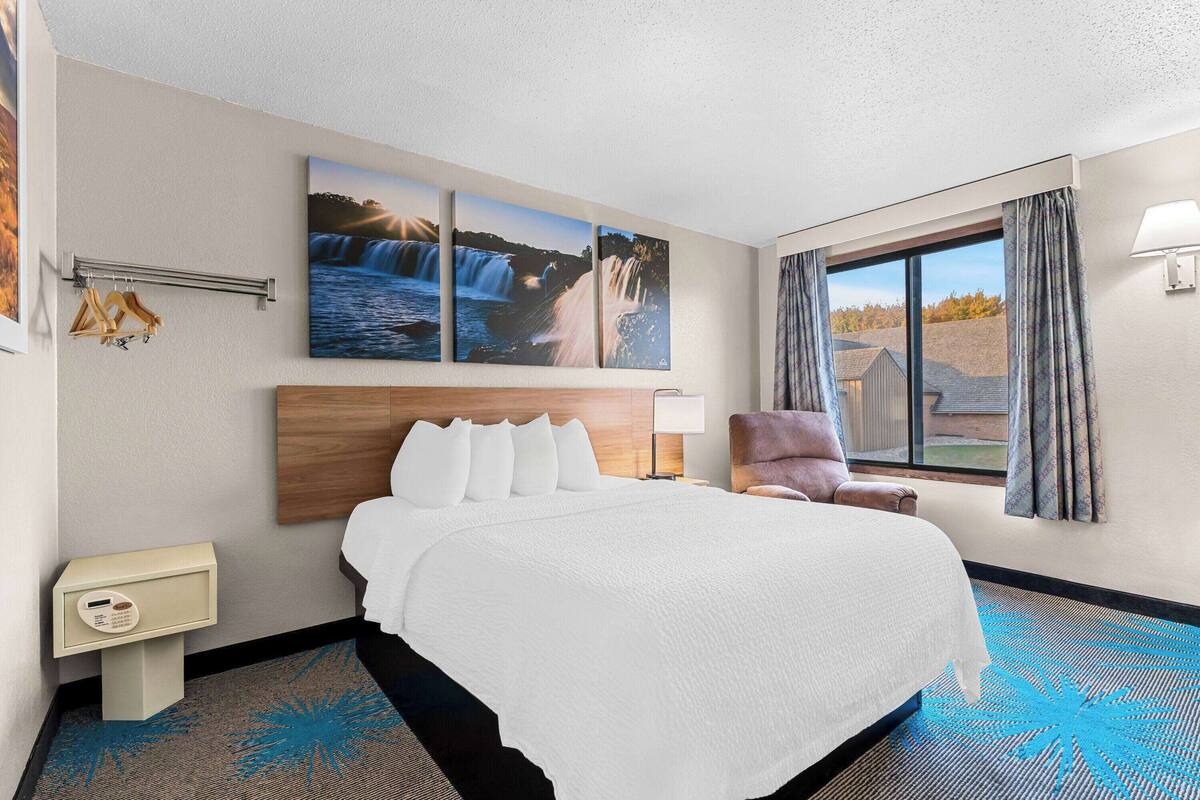 A hotel room with a large bed, a nightstand, a chair, a window with curtains, and three framed pictures on the wall.