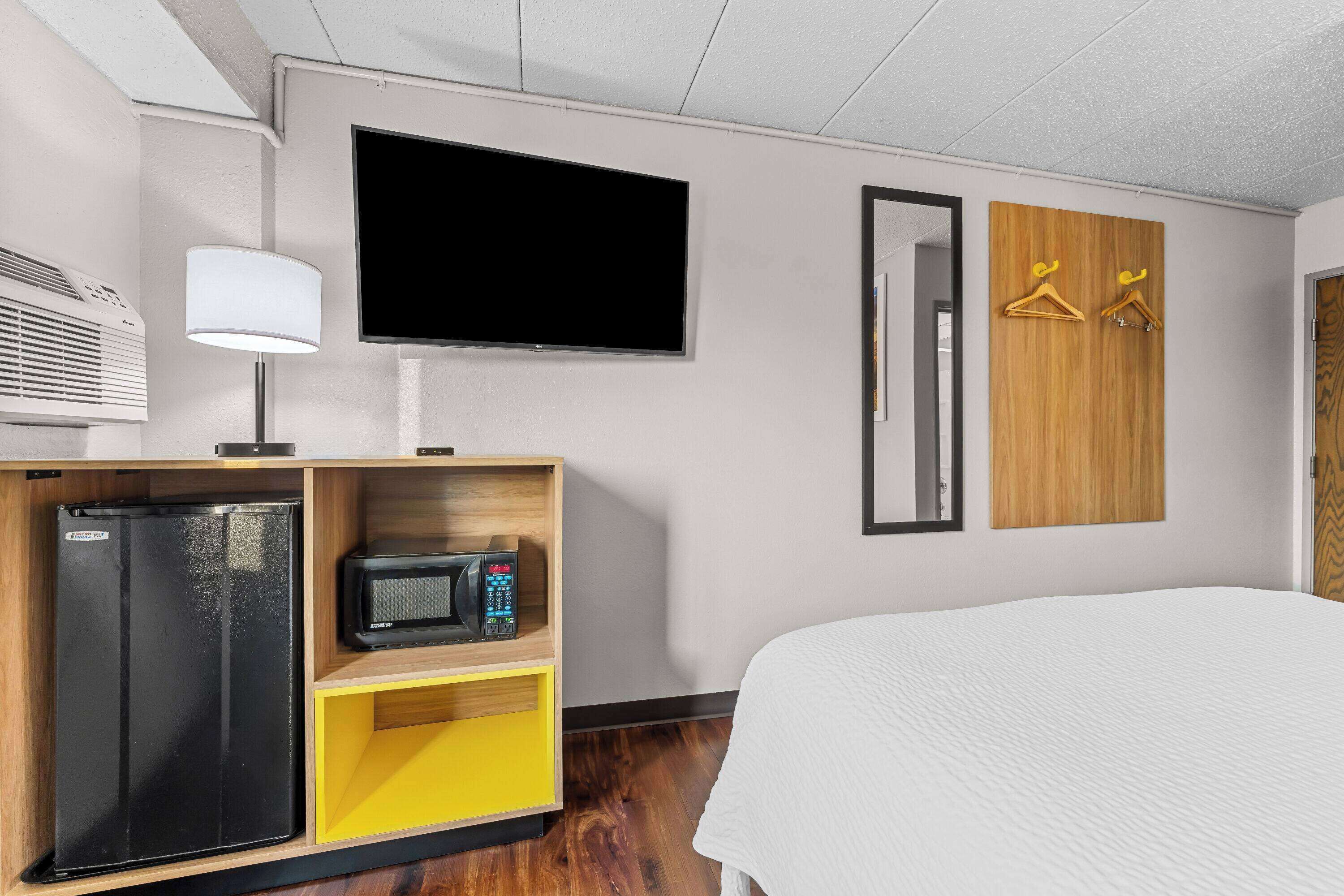 A hotel room with a bed, a TV, a mini-fridge, a microwave, a lamp, and wooden wardrobe with hangers.