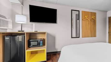 A hotel room with a bed, a TV, a mini-fridge, a microwave, a lamp, and wooden wardrobe with hangers.