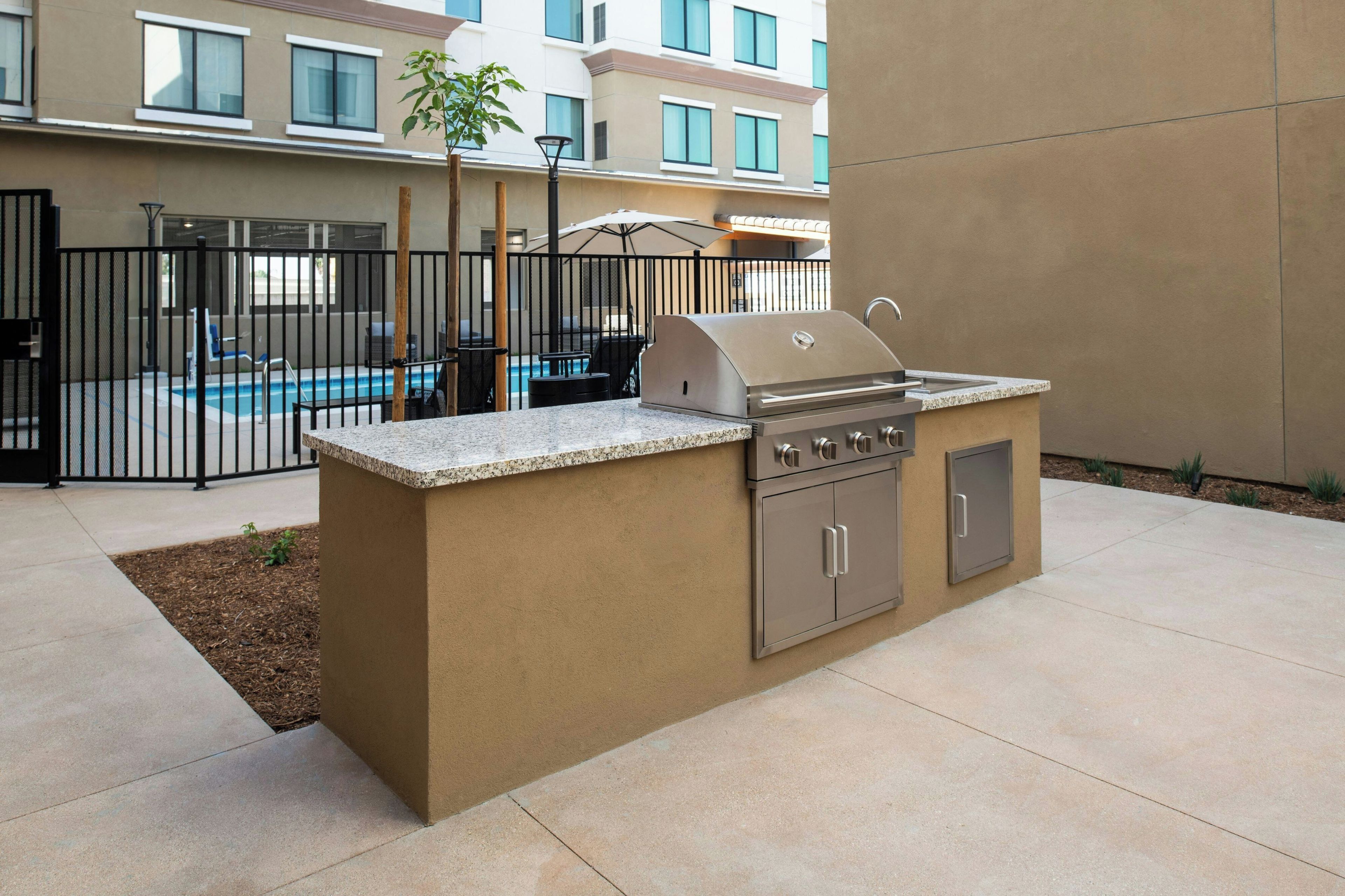 An outdoor kitchen area with a grill and sink, surrounded by a fence, with a pool and lounge chairs in the background.