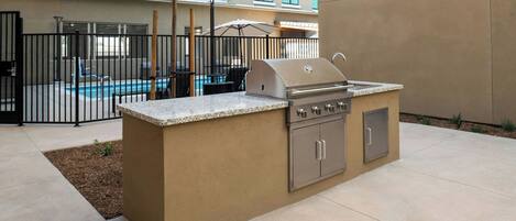 An outdoor kitchen area with a grill and sink, surrounded by a fence, with a pool and lounge chairs in the background.