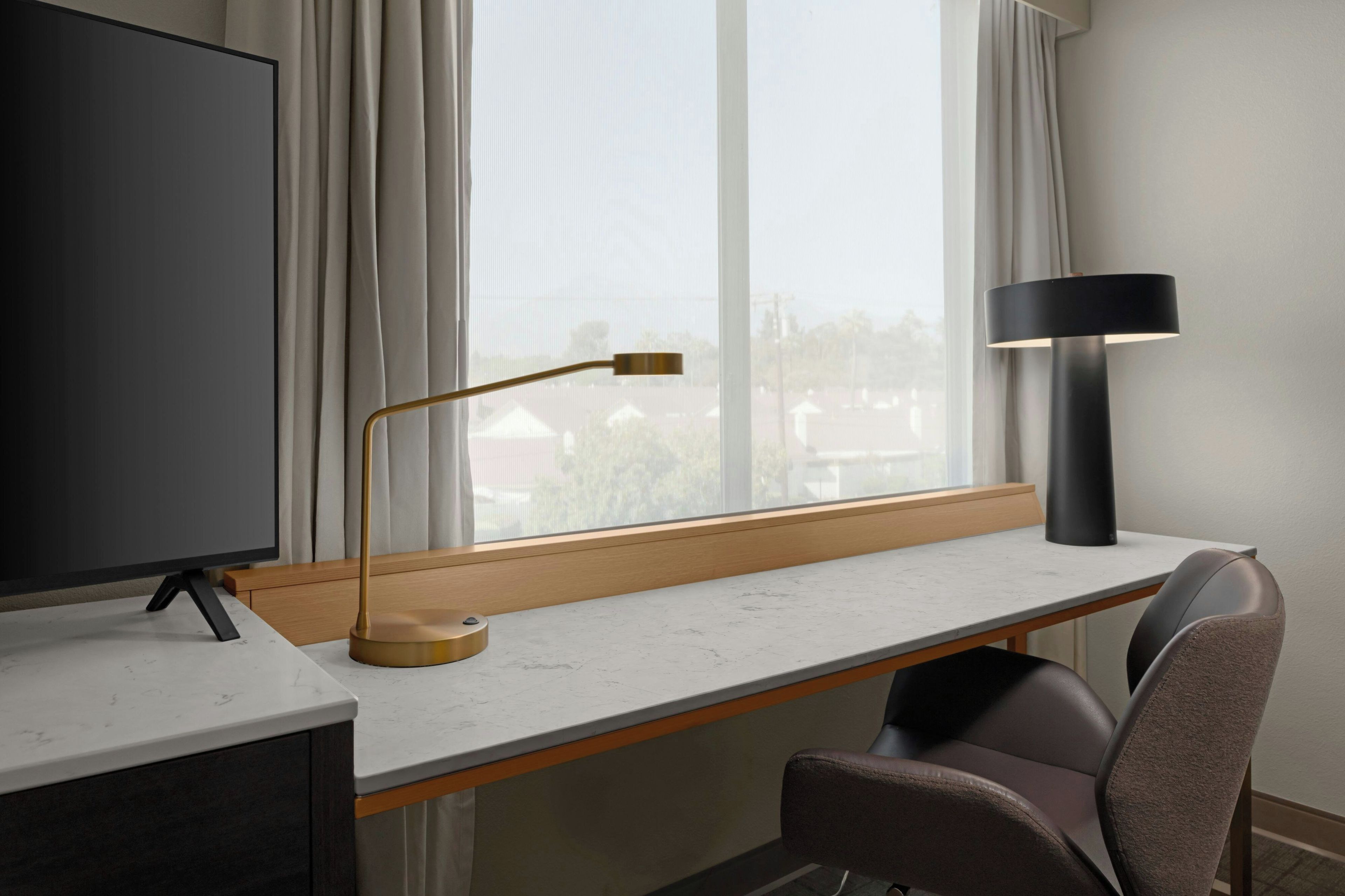 A modern hotel room with a large window, a desk with a flat-screen TV, a lamp, and a chair.