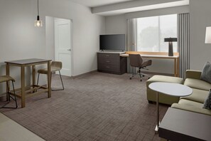 Ein Hotelzimmer mit Schreibtisch, zwei Stühlen, einer Couch, einem Fernseher und einem Fenster mit Vorhängen.