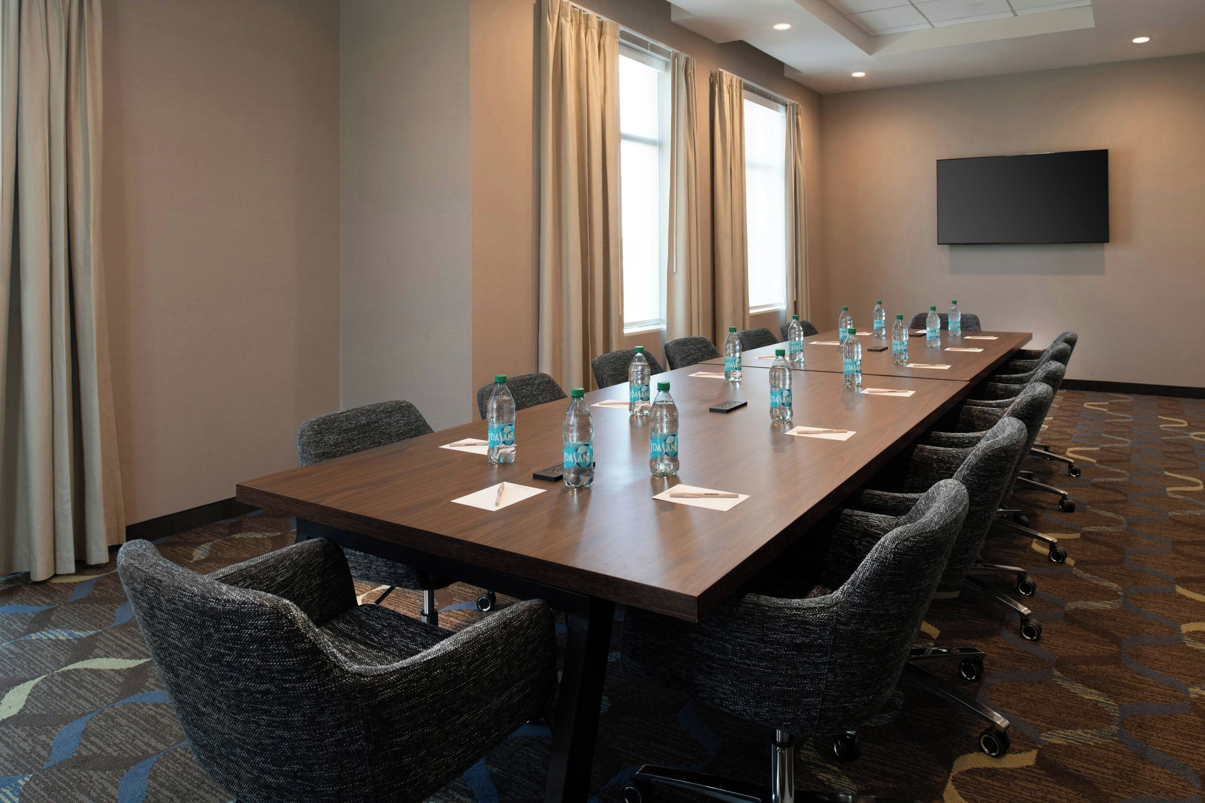 Un salón de conferencias con una larga mesa, sillas, botellas de agua y un televisor de pantalla plana en la pared.