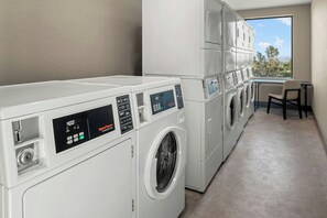 A laundry room with multiple washing machines and dryers, a window with a view of trees, and a small table with a chair. - Residence Inn by Marriott Claremont (Claremont)