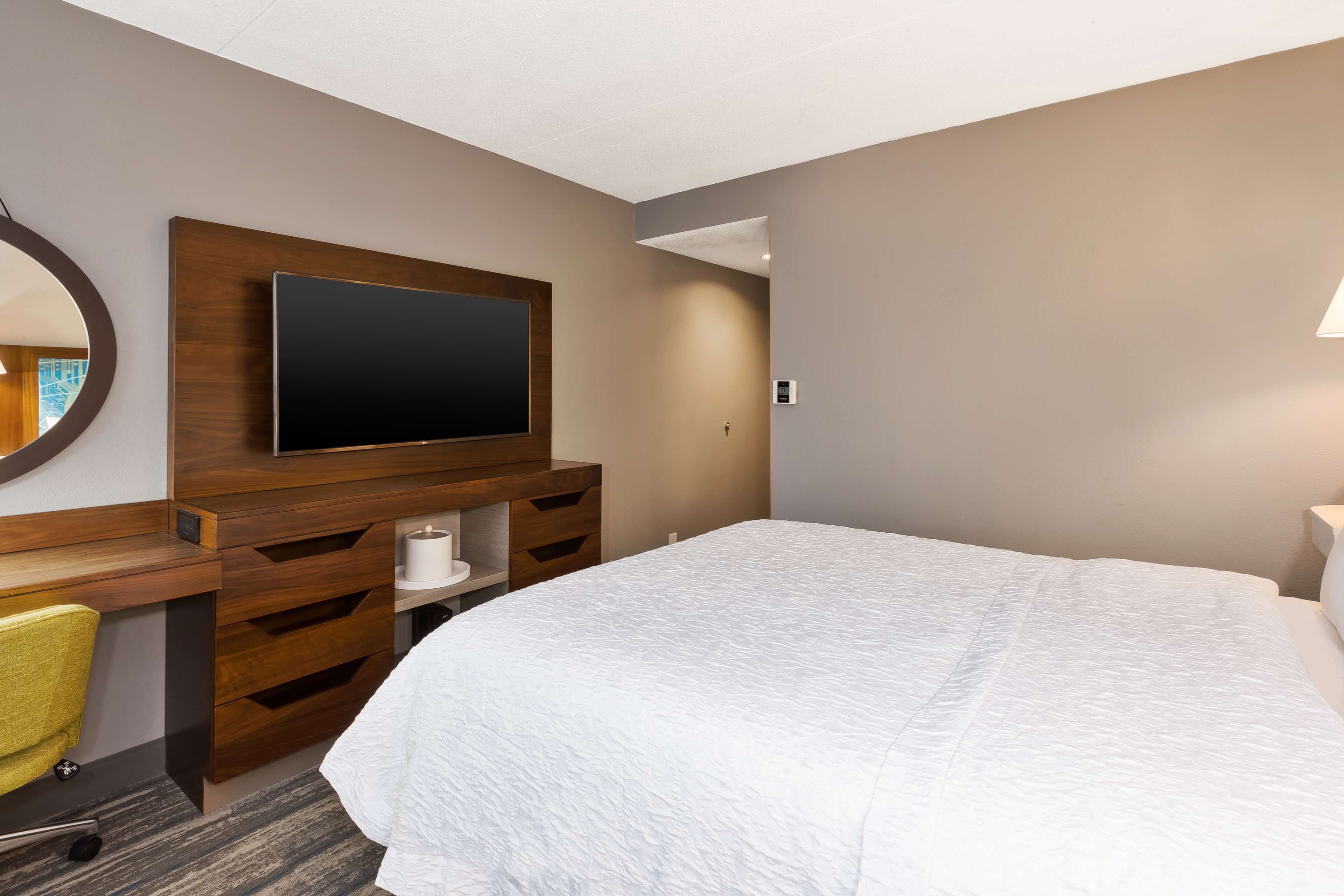 A hotel room with a large bed, a wooden desk with a flat-screen TV, a mirror, and a lamp.
