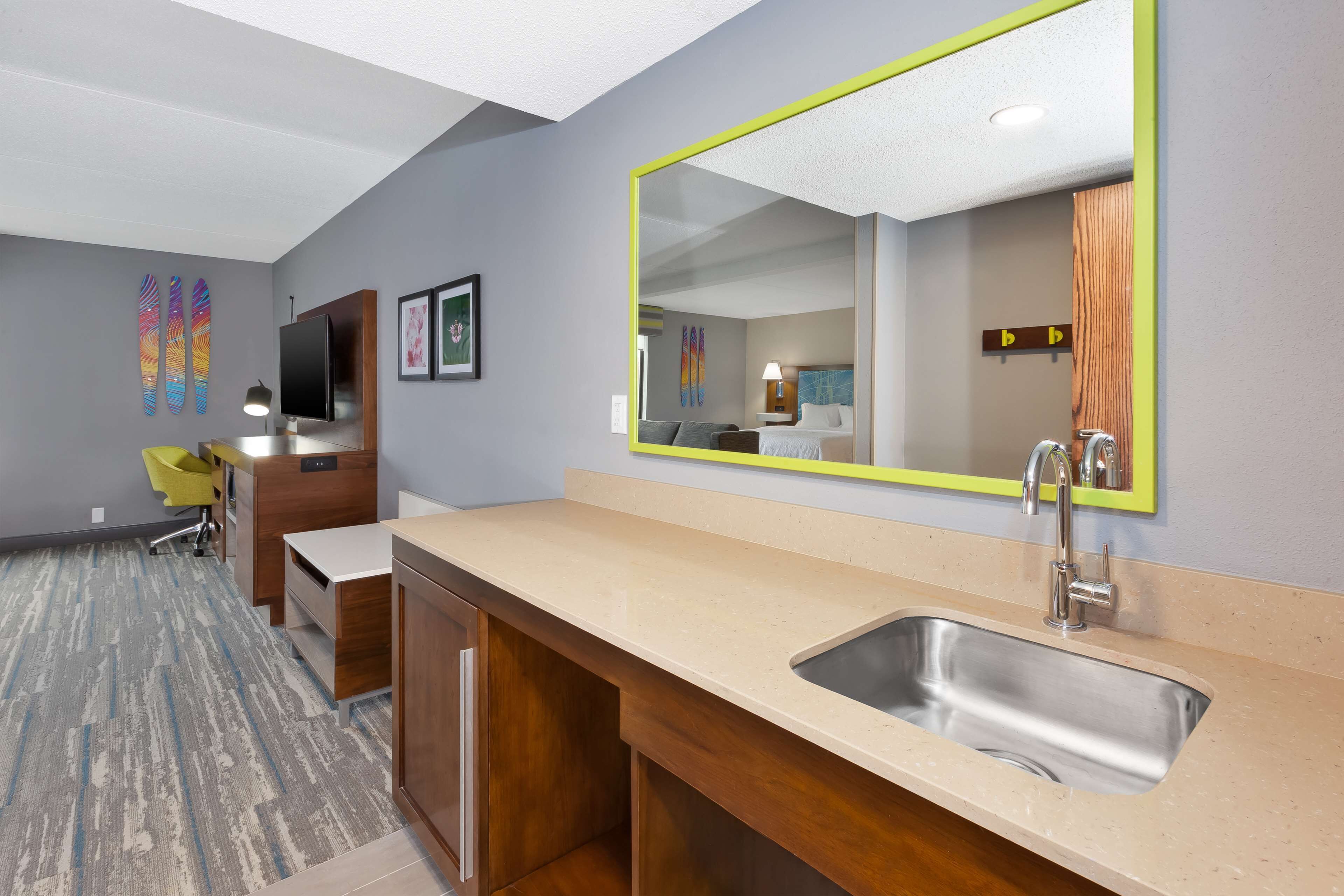 A modern hotel room with a large mirror, a sink, a wooden desk, and a chair.
