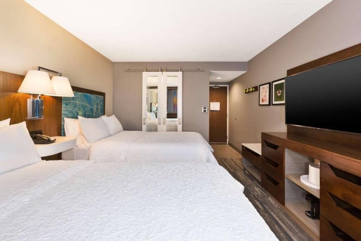 A hotel room with two beds, a desk with a lamp, a TV, and a large artwork on the wall.