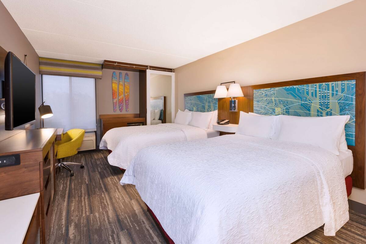A hotel room with two beds, a desk, a chair, and a TV.