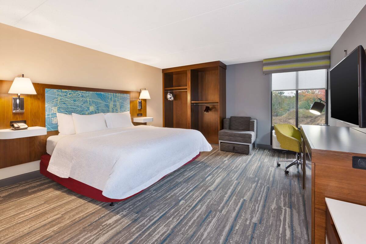 A hotel room with a large bed, a desk, a chair, a TV, and a window with a view of trees.