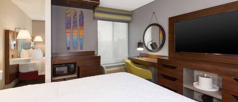 Ein Hotelzimmer mit einem großen Bett, einem Schreibtisch mit Stuhl, einem Fernseher, einer Mikrowelle und einem Badezimmerbereich mit Waschbecken und Spiegel.