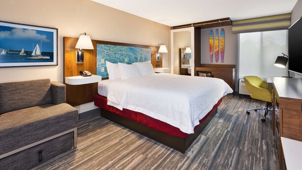 A hotel room with a large bed, a desk, a chair, a sofa, a painting, and a window with a view of the sea. - Hampton Inn Traverse City (Traverse City)