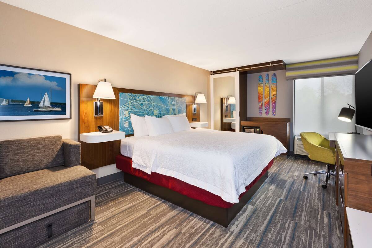 A hotel room with a large bed, a desk, a chair, a sofa, a painting, and a window with a view of the sea.