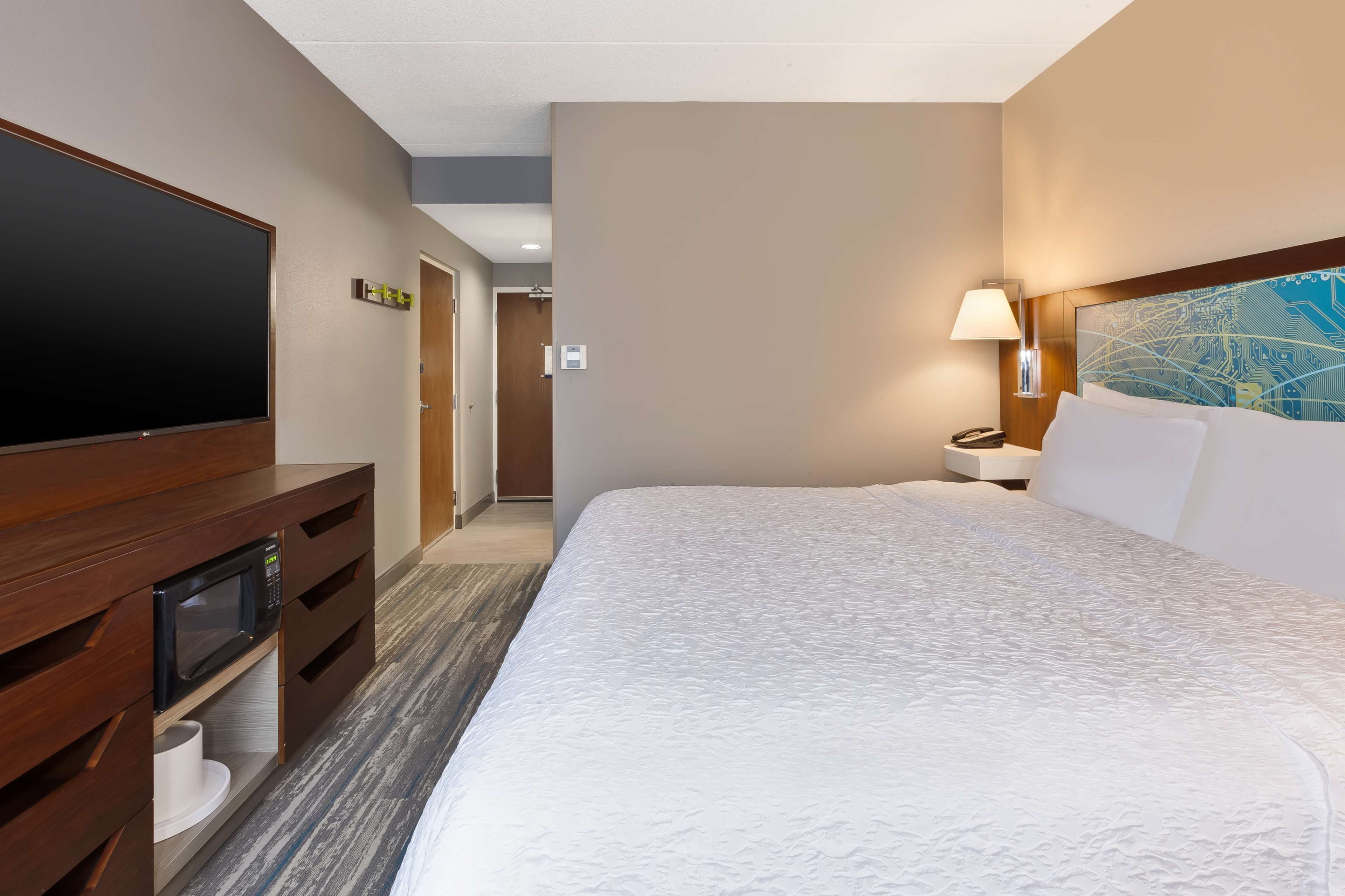 A hotel room with a large bed, a flat-screen TV, a wooden dresser, a nightstand with a lamp, and a wall art piece.