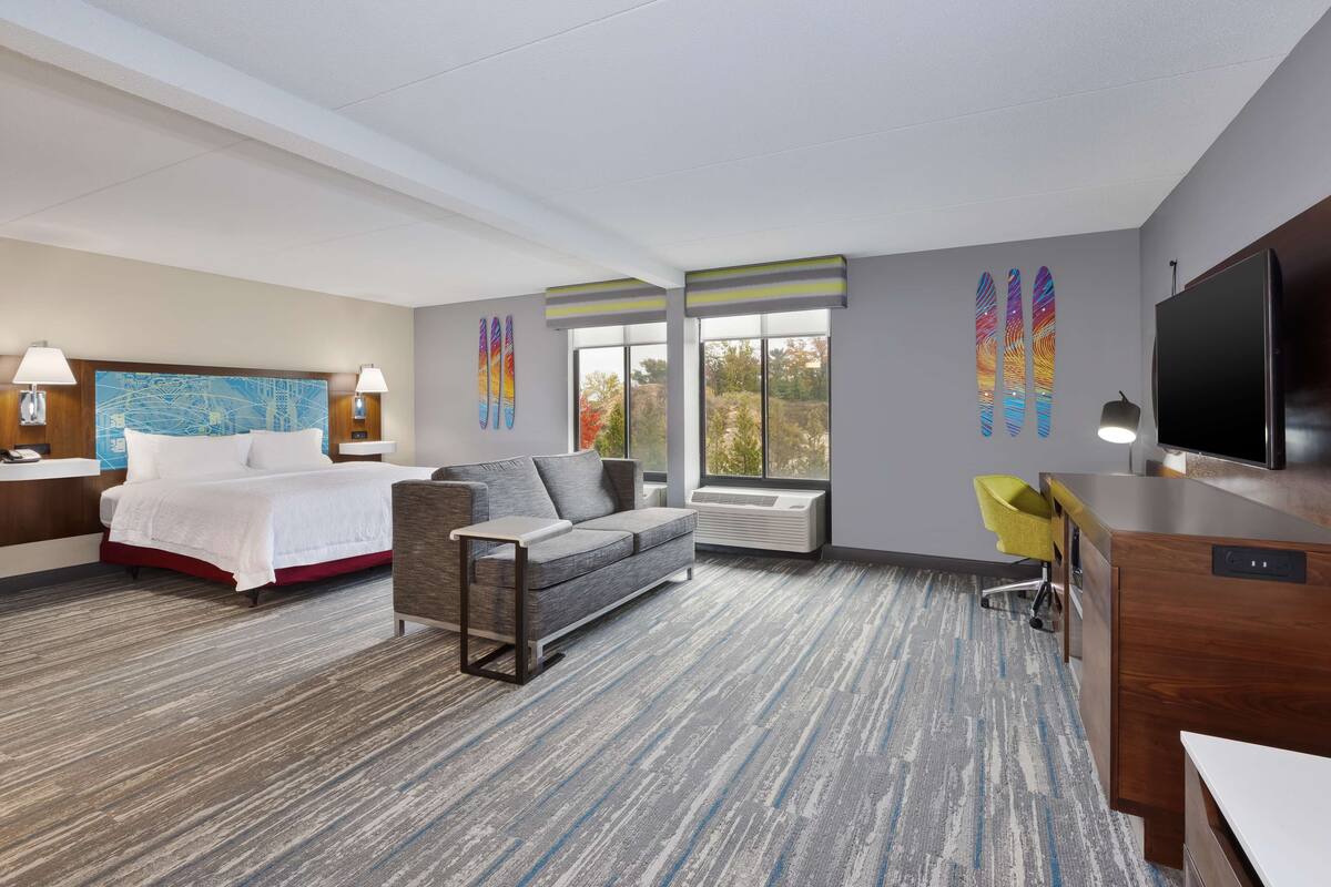 A hotel room with a bed, a sofa, a desk, a TV, and large windows.