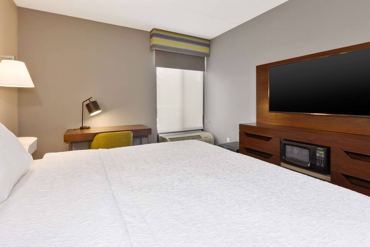 A hotel room with a bed, a desk with a lamp, a chair, a TV, and a microwave.