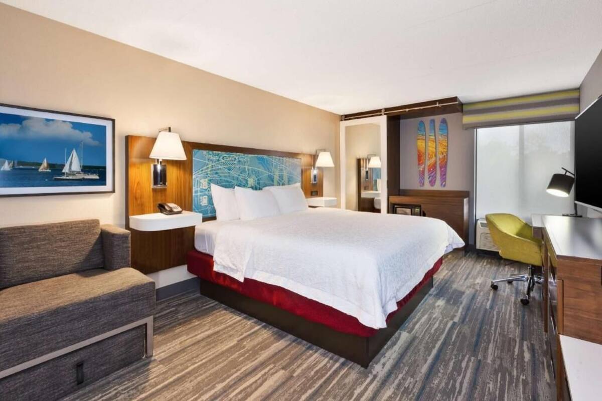 A hotel room with a large bed, a desk, a chair, a sofa, a painting, a map, and a television.