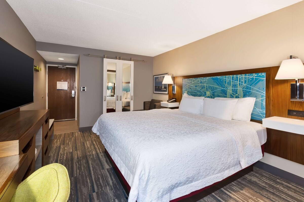 A hotel room with a large bed, a desk, a chair, a TV, and a bathroom with a mirror and towel rack.
