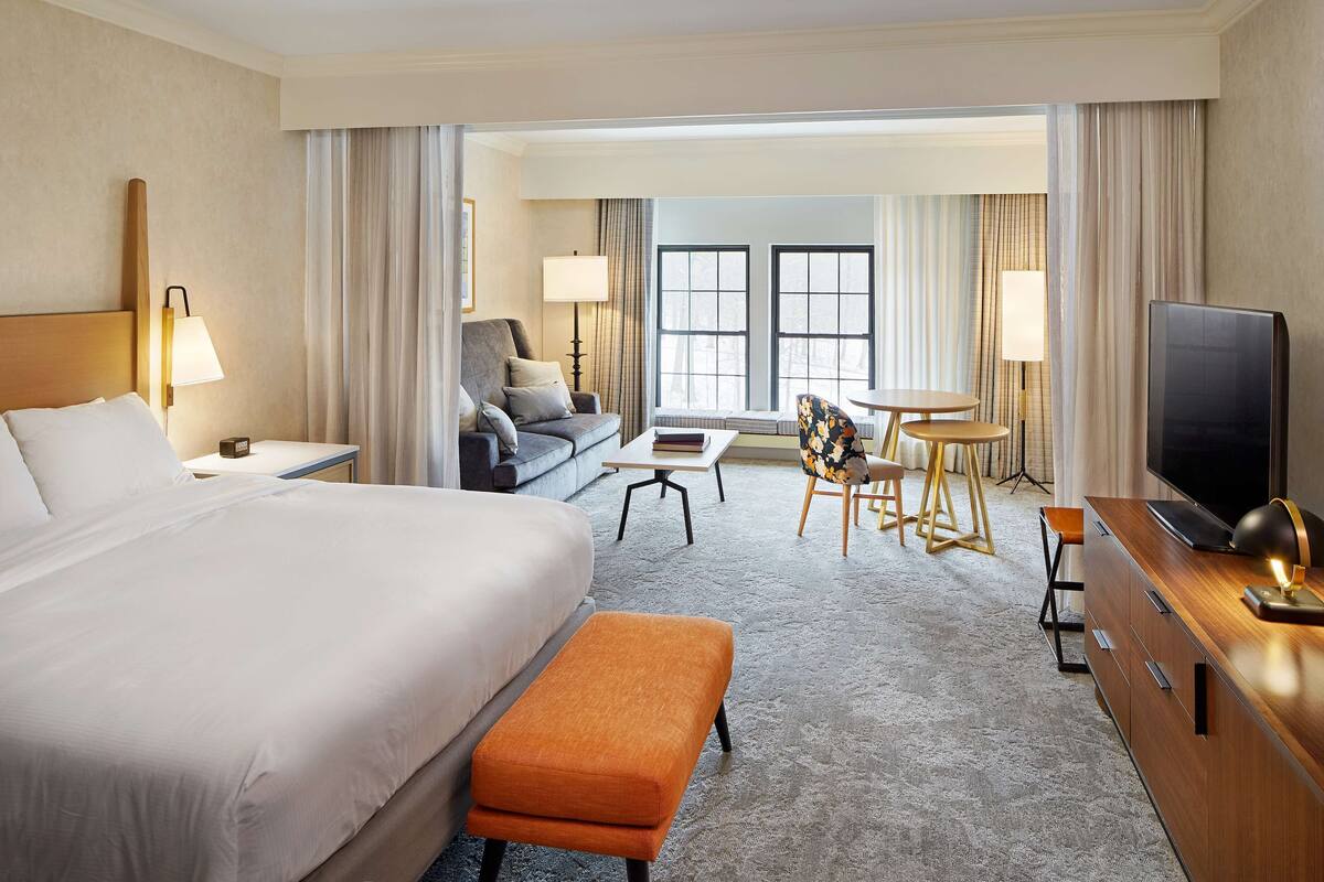 A hotel room with a large bed, an orange bench, a TV, a desk, and a chair.
