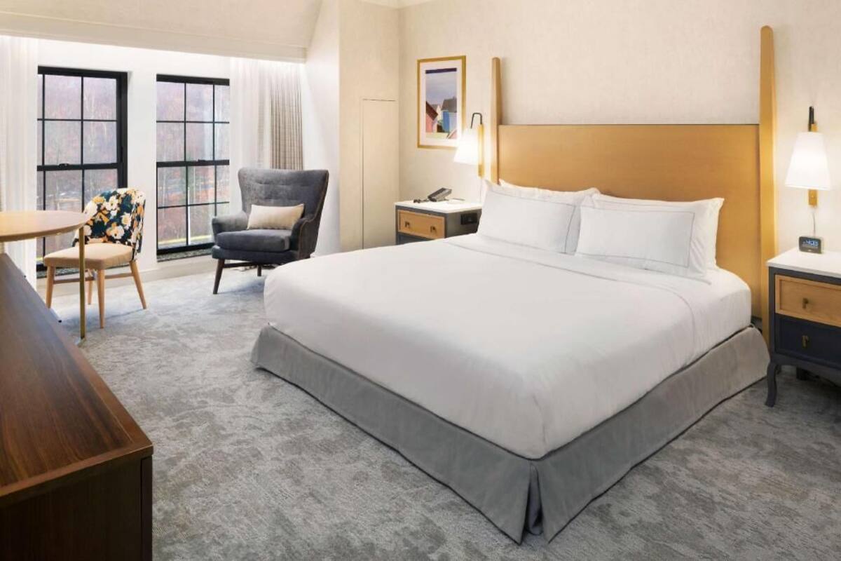 A hotel room with a large bed, a desk, a chair, and a nightstand with a lamp.