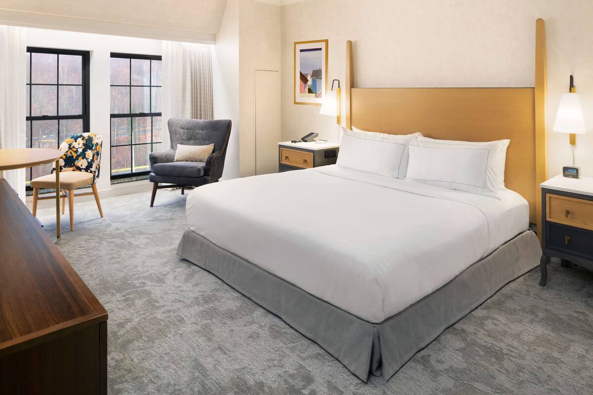 A hotel room with a large bed, a desk, a chair, and a nightstand with a lamp.