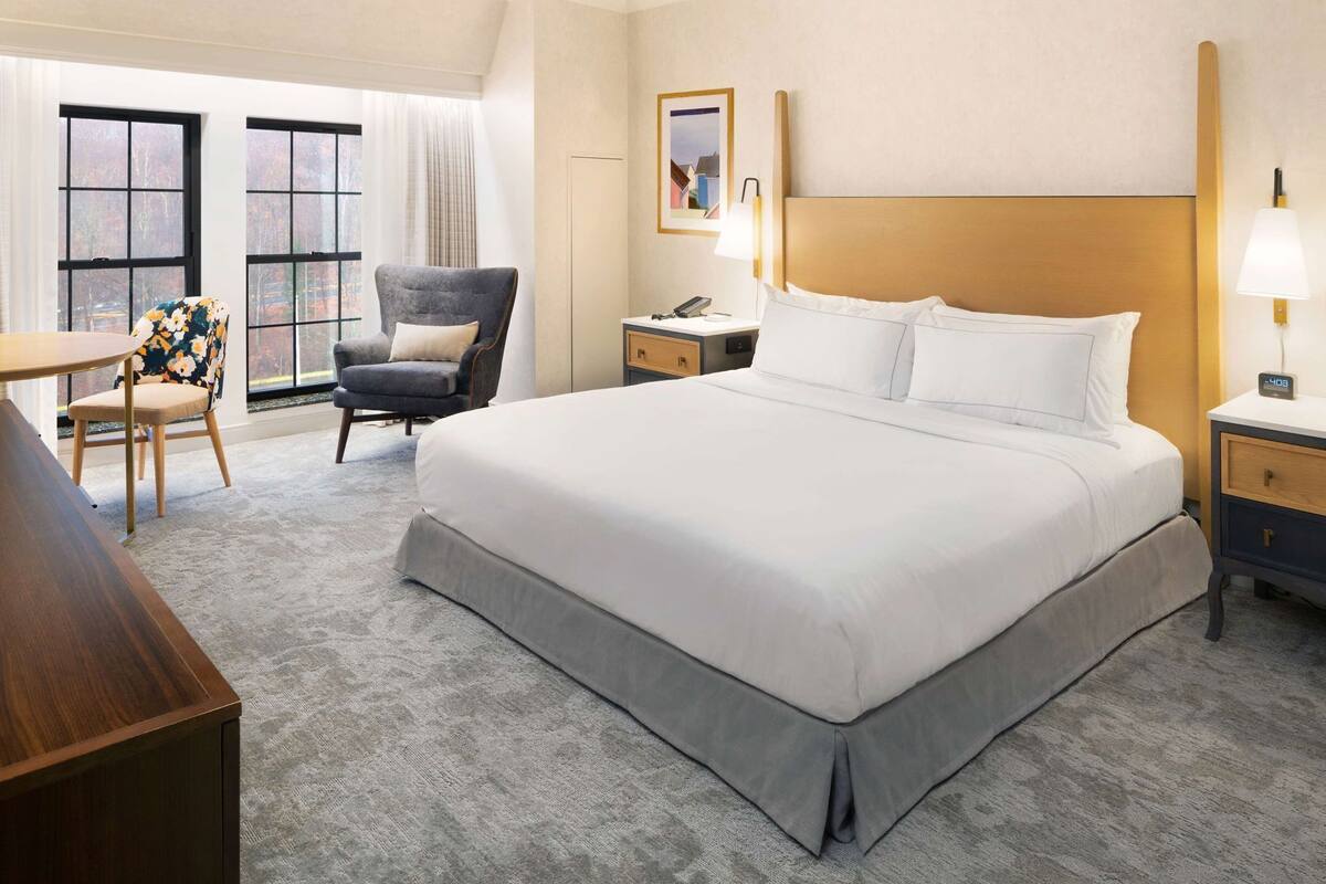 A hotel room with a large bed, a desk, a chair, and a nightstand with a lamp.