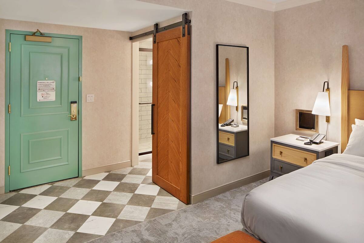 A hotel room with a checkered floor, a green door, a wooden sliding door, a bedside table with a lamp, a mirror, and a bed.