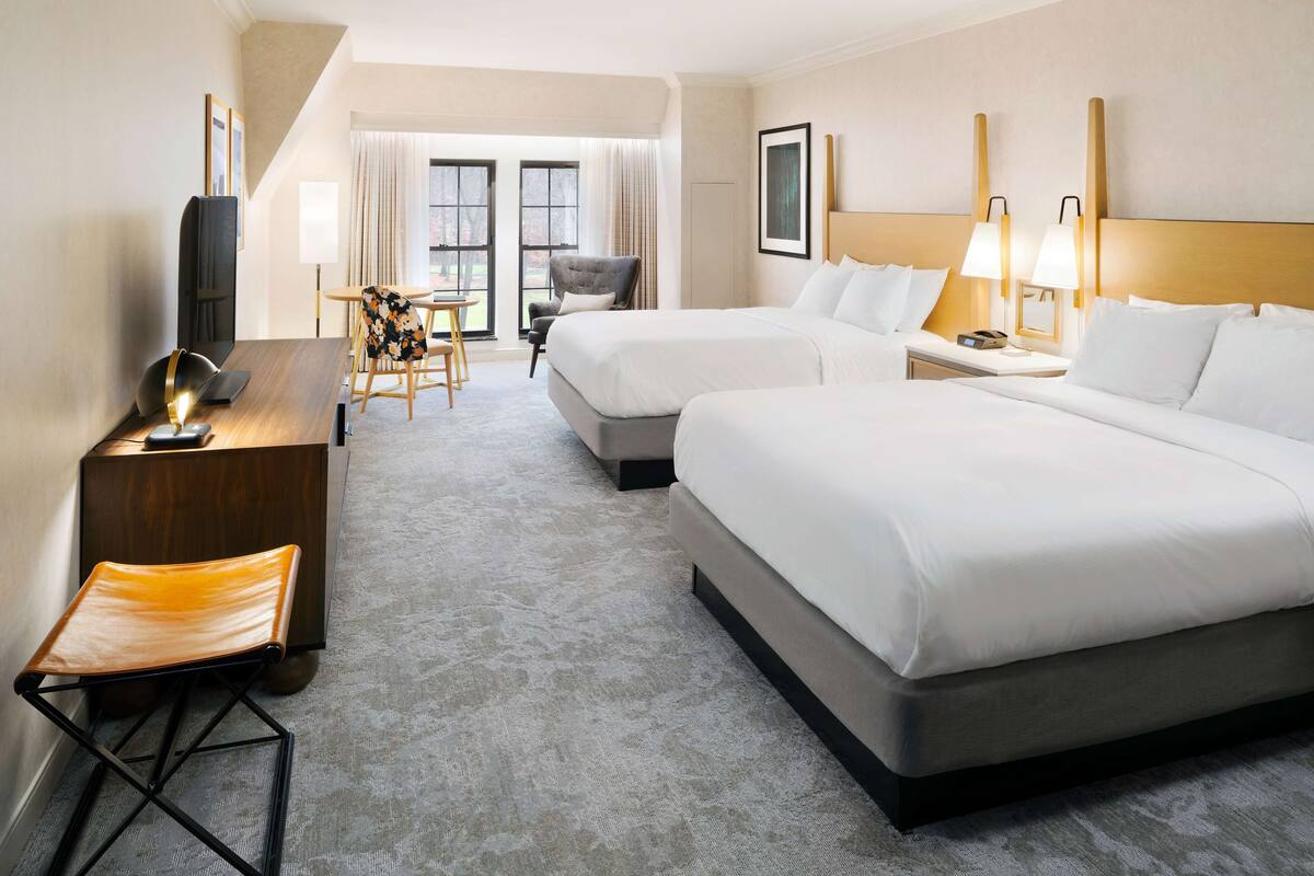 A hotel room with two beds, a desk with a chair, a TV, and a window with curtains.
