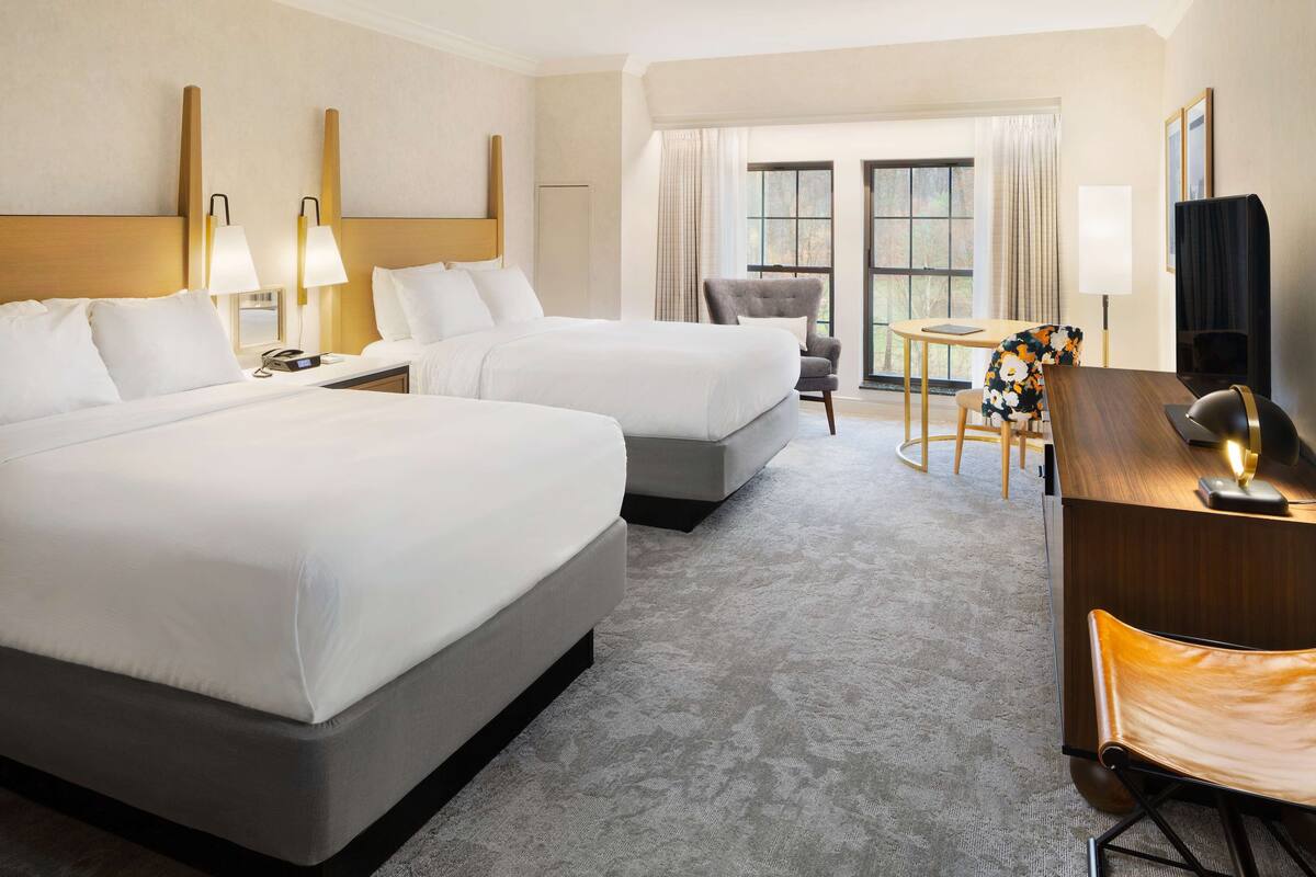 A hotel room with two beds, a desk, a chair, and a TV.