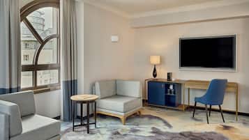 A hotel room with a sofa, a small round table, a desk with a chair, a TV, and a view of a building through a window.