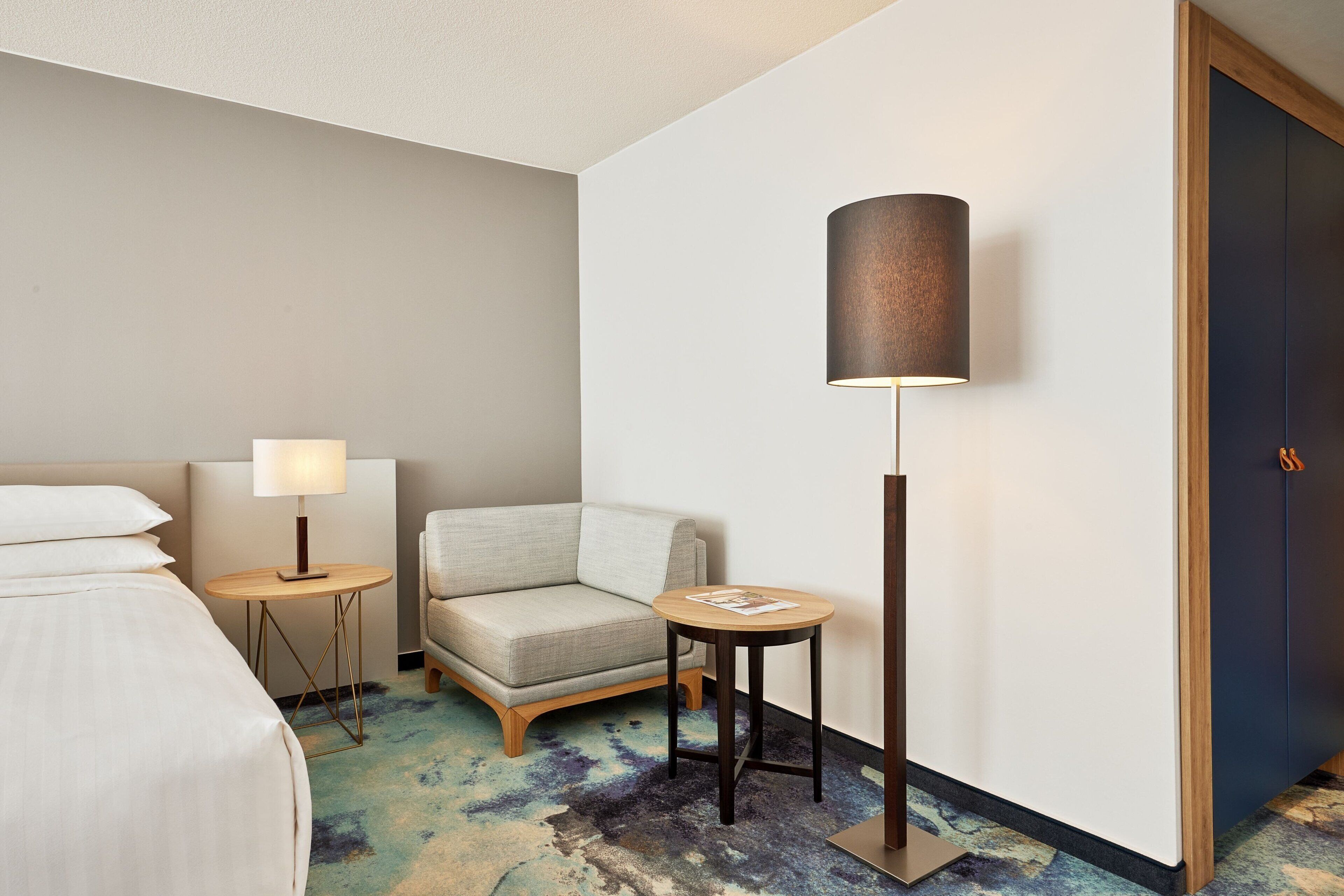 Une chambre d’hôtel avec un lit, une chaise, une petite table, une lampe et une porte.