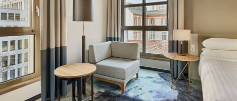 Habitación de hotel con sofá, una mesa redonda, lámpara, cama y vista a edificios a través de ventanas amplias.