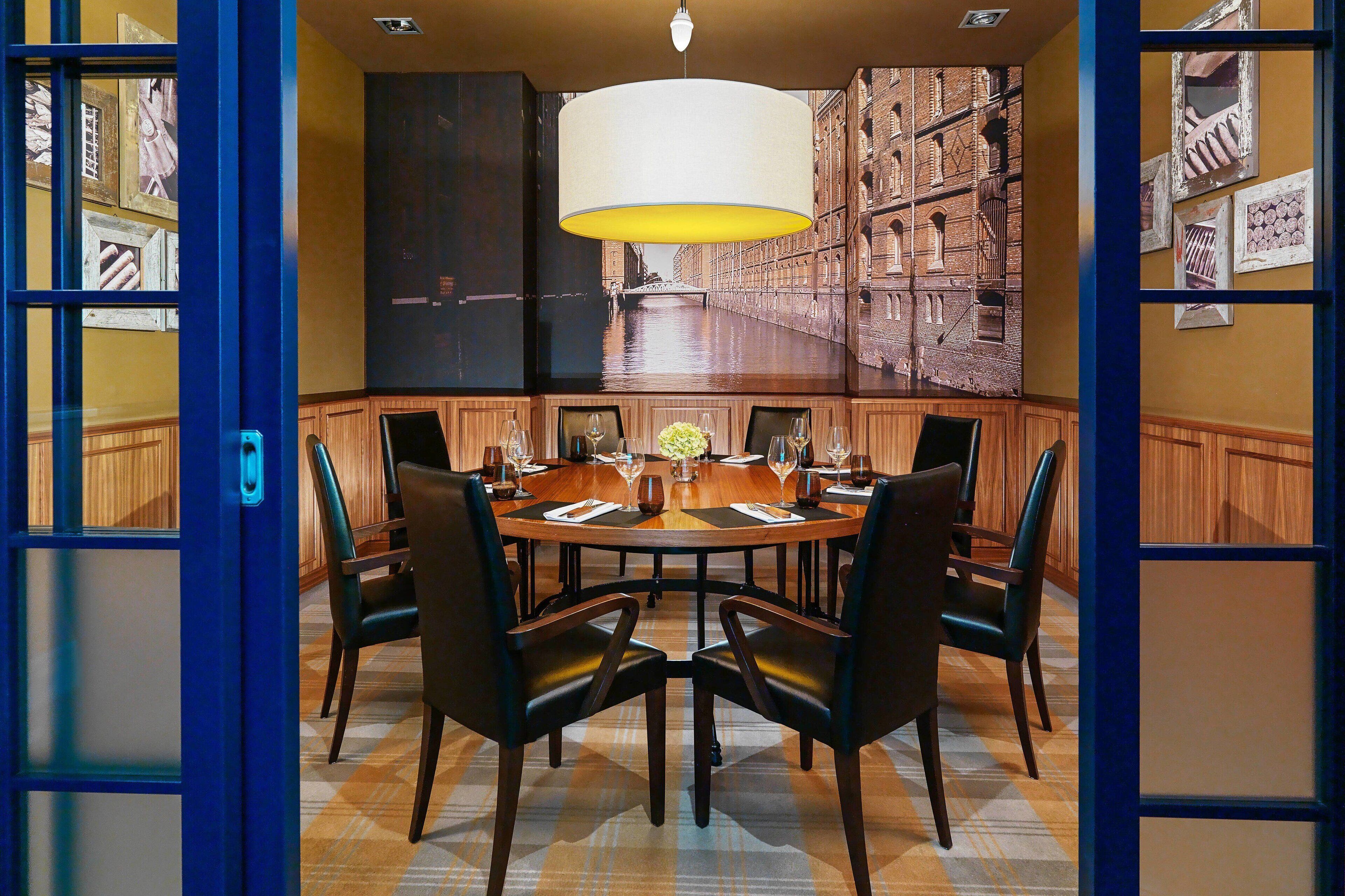 A dining area with a round table, six chairs, and a large painting of a cityscape on the wall.