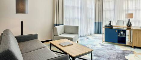 Una habitación de hotel moderna con un sofá gris, una mesa de centro de madera, un escritorio con lámpara y un ventanal con cortinas translúcidas.