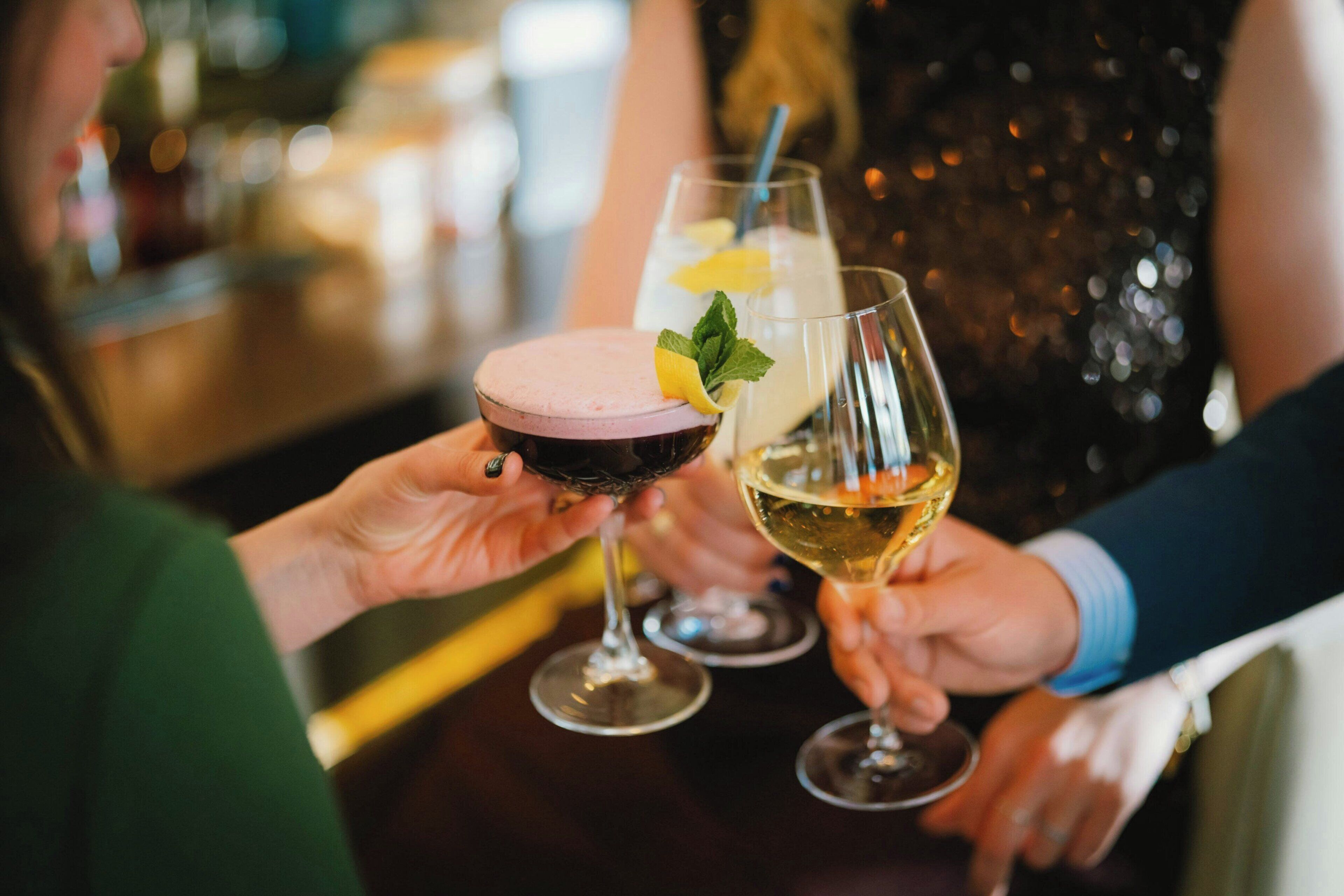Dos personas sosteniendo vasos con bebidas, uno con un trago rosado y el otro con un trago transparente, en un bar.
