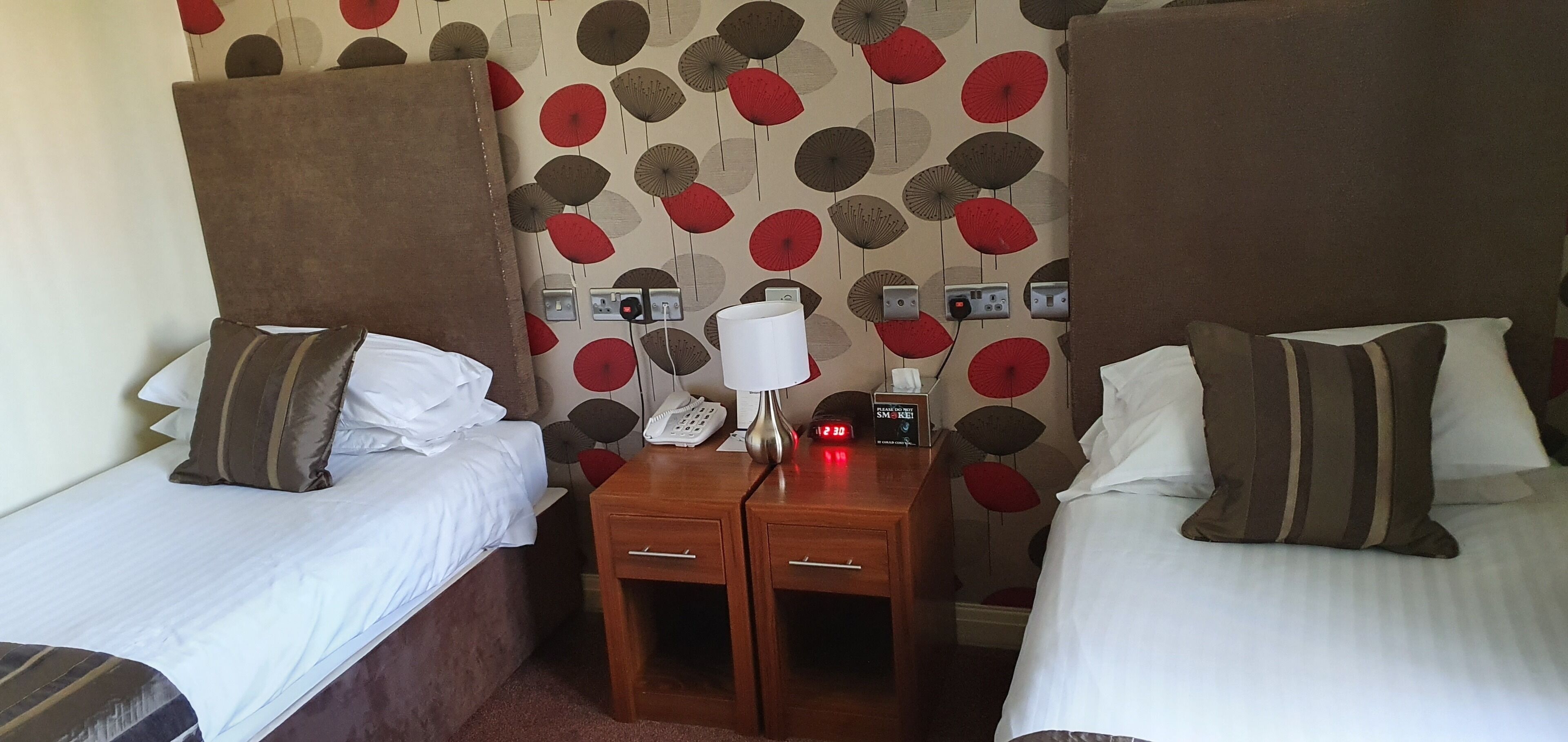 A hotel room with two beds, a nightstand with a lamp, a clock, and a phone.