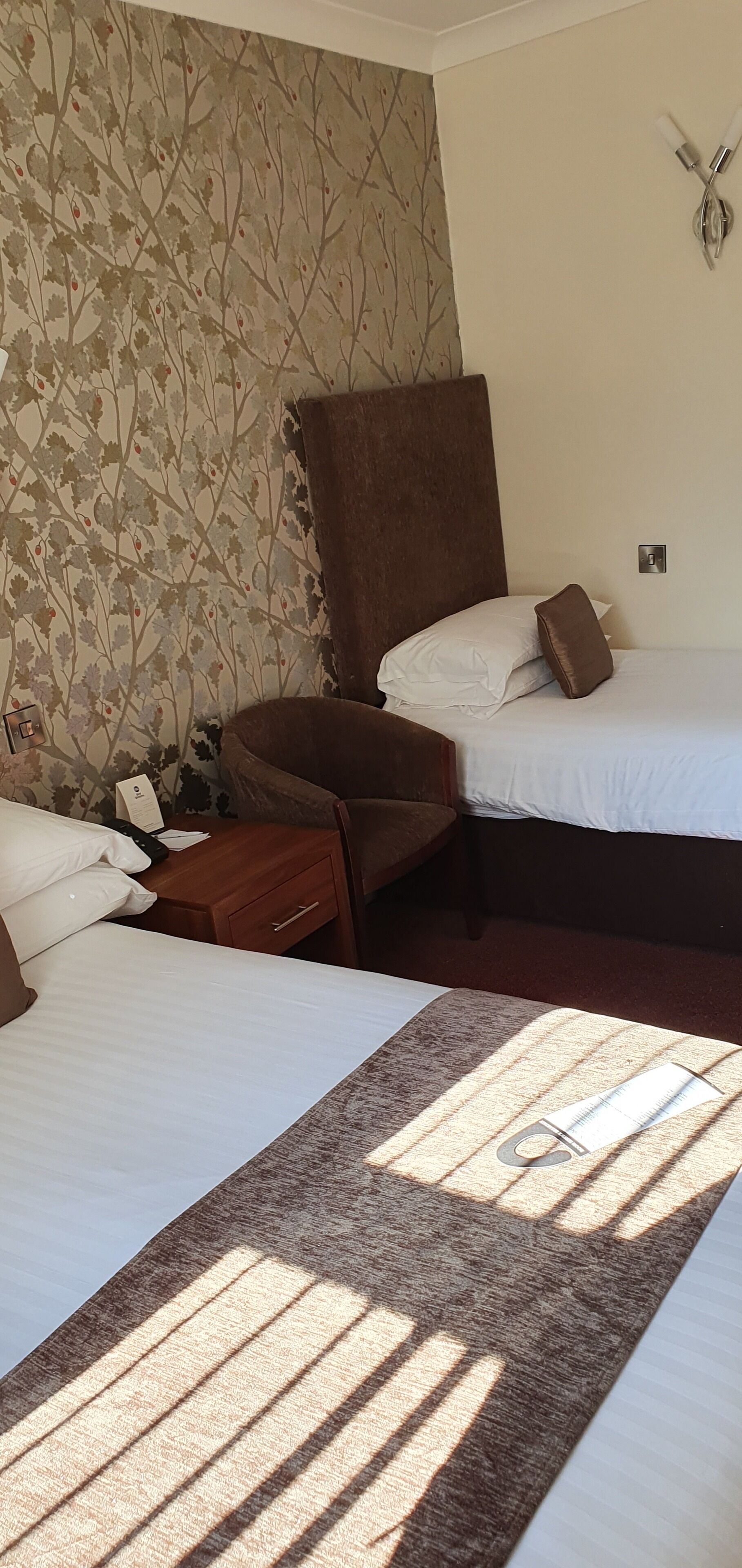 A hotel room with two beds, a brown armchair, a wooden nightstand, and a patterned wallpaper.