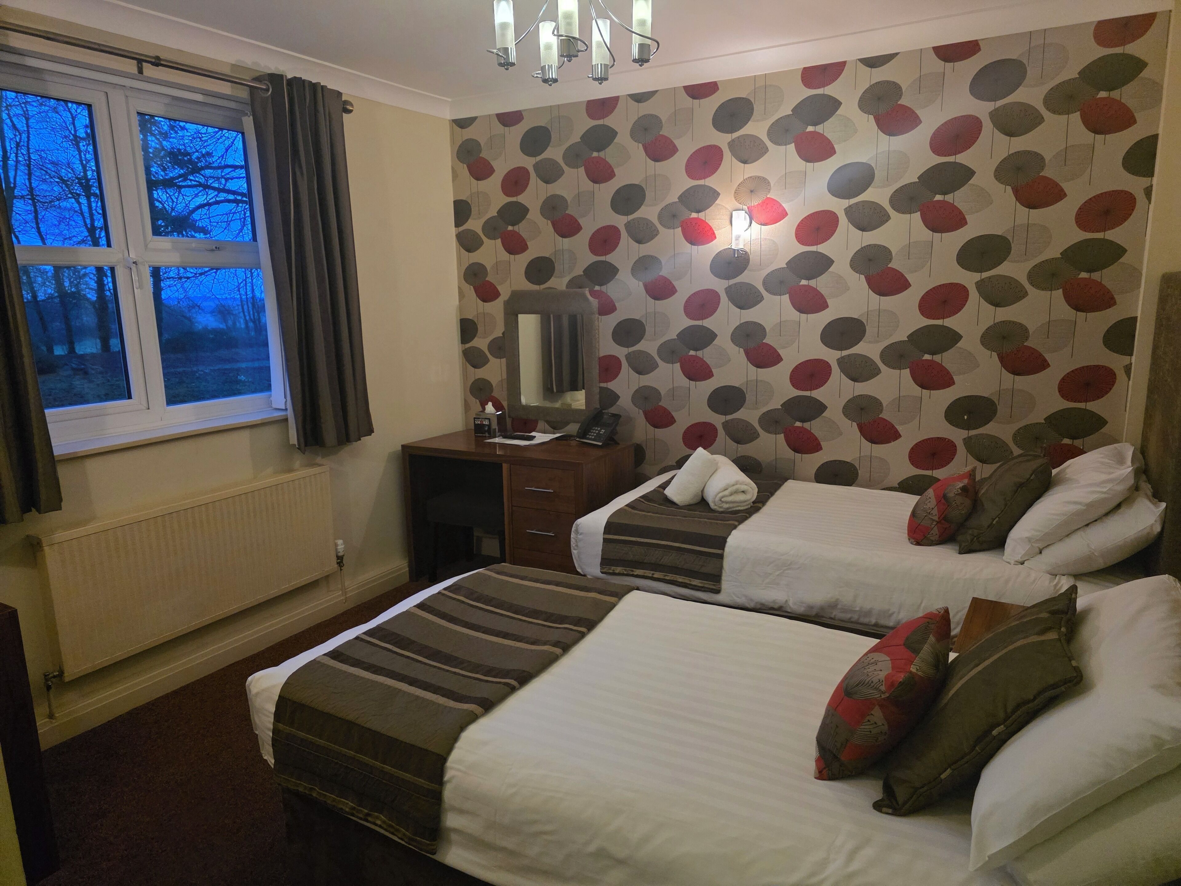 Ein Hotelzimmer mit zwei Betten, einem Schreibtisch, einem Sessel, einer Heizung, einem Fenster mit Vorhängen und einer Wand mit Muster.