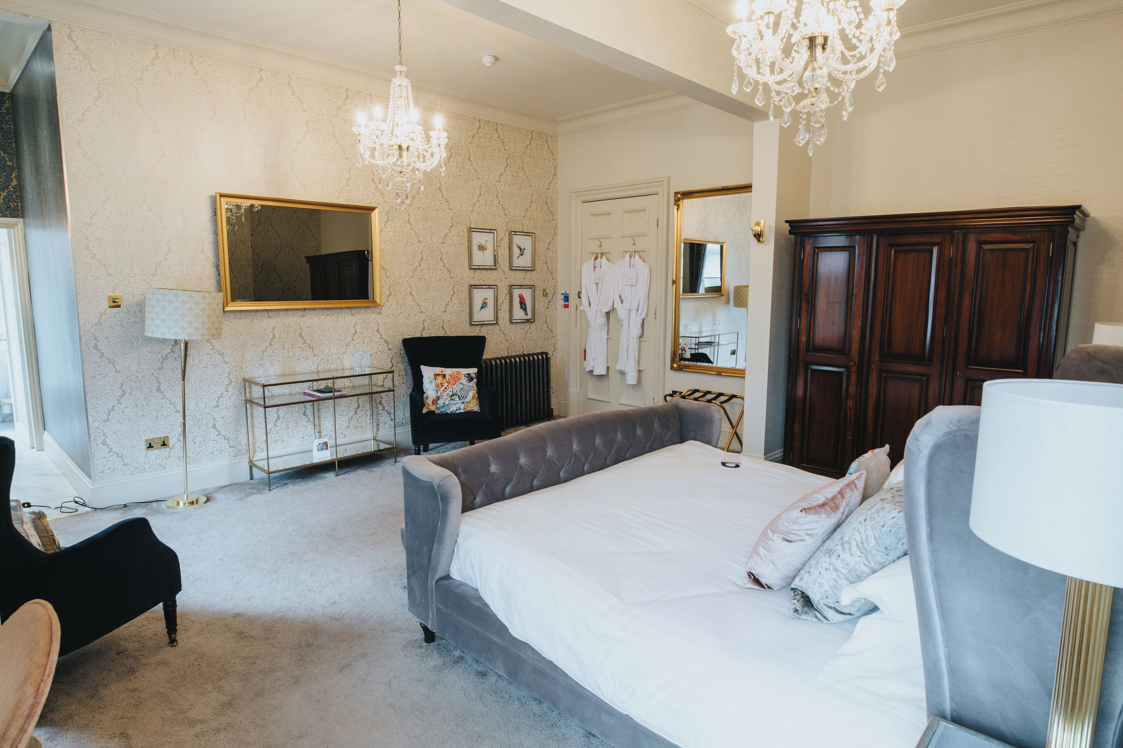 A spacious bedroom with a large bed, a sitting area with a chair, a vanity with a mirror, and a wardrobe.