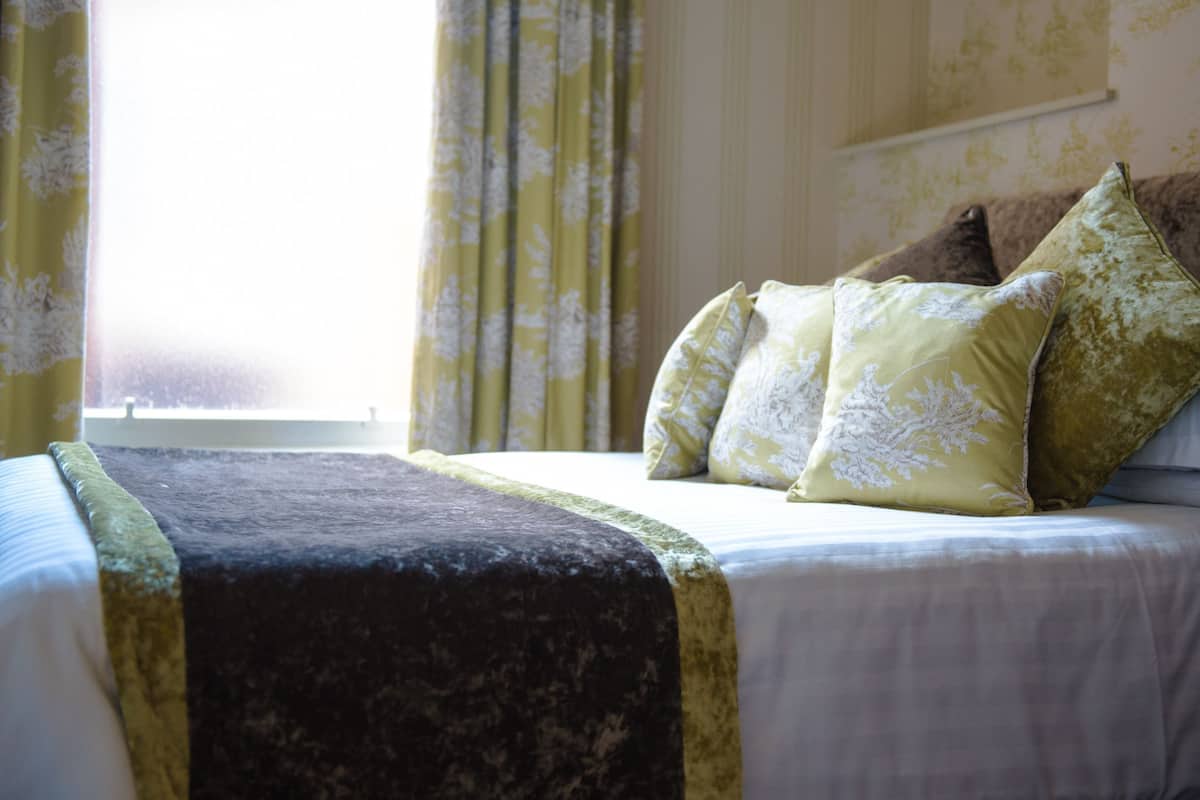 A neatly made bed with a dark blanket and multiple pillows, a window with curtains, and a wall with a floral pattern.