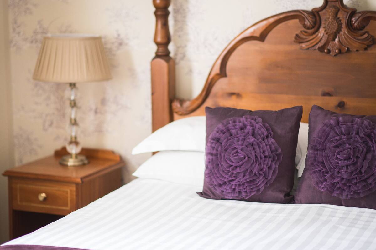 A neatly made bed with a wooden headboard, two purple pillows, and a bedside table with a lamp.