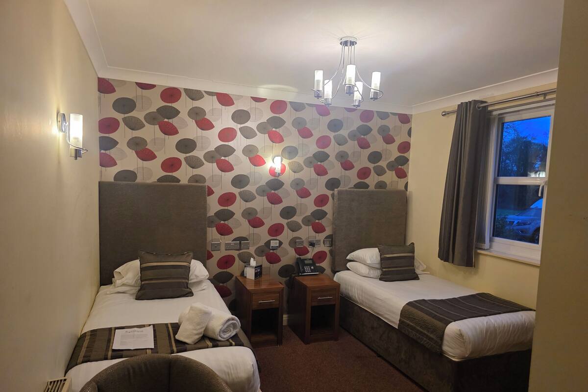 A hotel room with two beds, a desk, a chair, a radiator, and a window with curtains.